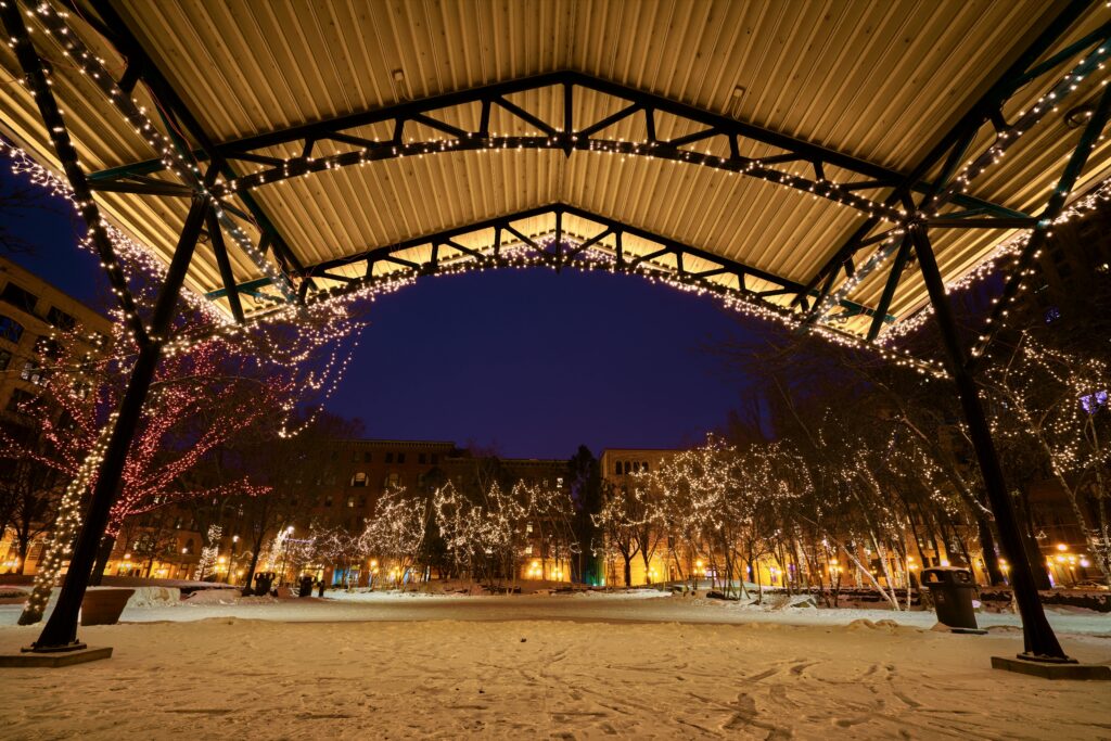 Snowy city park at dusk under a light-strung pavilion, trees wrapped in twinkle lights creating a festive winter scene.
